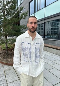 Vit linneskjorta med blå broderade mönster på framsidan, med krage och långa ärmar. Buren med ljusa cargo-shorts.