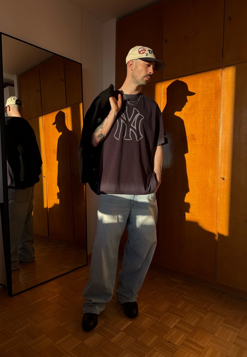 Hombre con gorra, camiseta oscura de NY y vaqueros claros sostiene una chaqueta sobre el hombro, de pie junto a un armario de madera con sombra reflejada en la habitación.