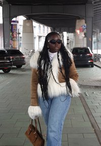 Chaqueta de piel sintética marrón con detalles de piel blanca, suéter de cuello alto de color claro, vaqueros acampanados azules y bolso marrón, bajo un paso elevado.