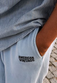 Prugasta modra in bela majica s kratkimi rokavi nad svetlo modrimi trenirkami, ki imajo na levem žepu črn logotip "American Vintage".