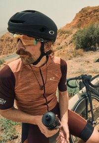 Jalgrattasõiduriietus, millel on punakaspruun lühikeste varrukatega jersy, must kiiver ja peegelprillid. Hoides musta joogipudelit jalgratta lähedal.