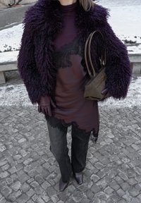 Personne portant une veste en fausse fourrure violette, un haut transparent orné de dentelle, un pantalon noir, des gants et des chaussures violets, tenant un sac à main vert olive sur un trottoir enneigé.