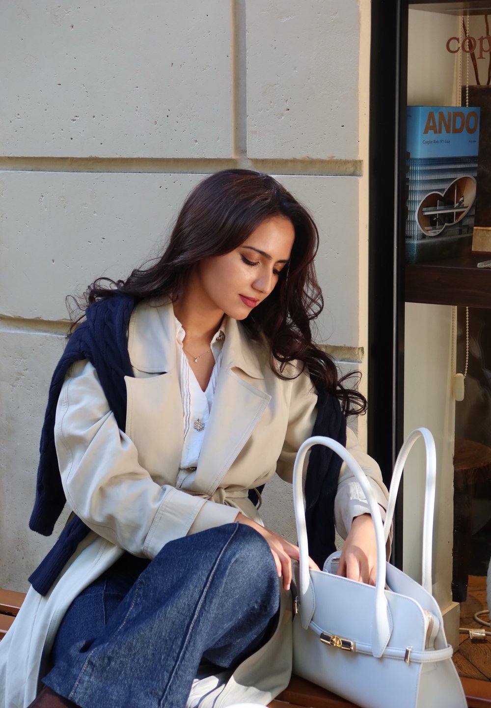 Beiger Trenchcoat über einem marineblauen Pullover, blauer Denim und eine weiße Handtasche mit zwei Griffen und goldenen Beschlägen, vor einer hellen Wand.