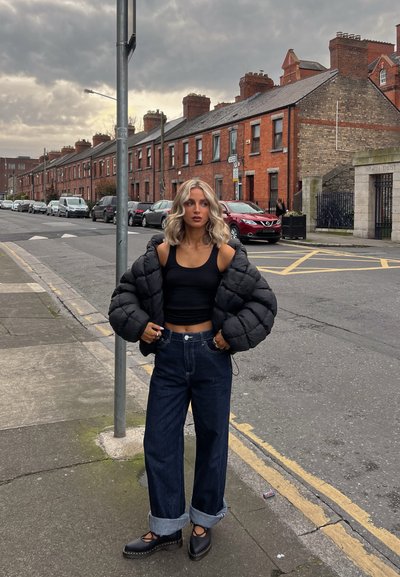 Chaqueta de plumón negra sobre un top corto negro, combinada con jeans de pierna ancha. Zapatos de plataforma negros con detalles recortados. Entorno urbano.