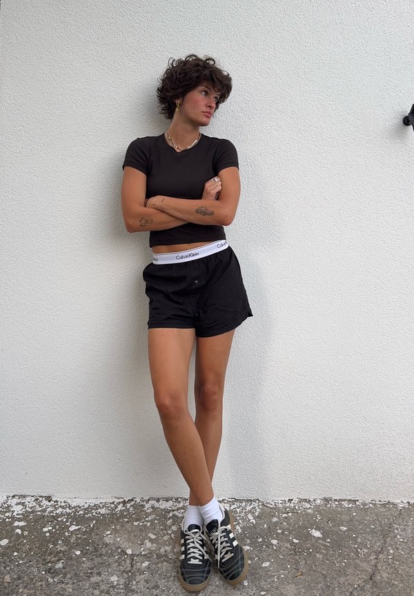 Person mit kurzen lockigen Haaren, die ein schwarzes Shirt und Shorts, weiße Socken und schwarze Turnschuhe trägt, steht mit verschränkten Armen vor einer weißen Wand.