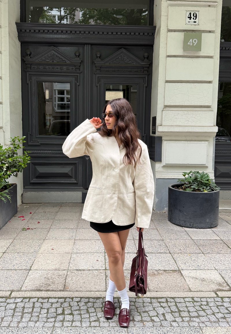 Mujer con cabello largo que lleva gafas de sol, blazer beige de gran tamaño, falda negra, calcetines blancos, zapatos burdeos y sostiene un bolso burdeos mientras está de pie junto a una puerta.