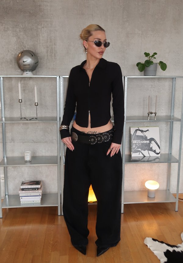 Žena v čiernom top s na zips, nohavice s širokými nohavicami, opasok s cvokmi, slnečné okuliare, stojaca v moderne zariadenej miestnosti s poličkami, rastlinami a knihami.