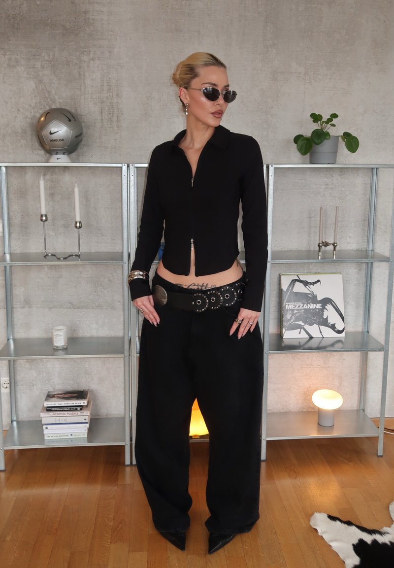 Femme portant un haut noir à fermeture éclair, pantalon à jambes larges, ceinture cloutée, lunettes de soleil, se tenant dans une pièce moderne avec des étagères, des plantes et des livres.