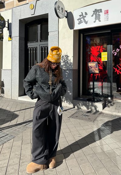 Chaqueta corta negra de lana, pantalones de pierna ancha grises, botas color marrón. Gorra amarilla con un diseño, collares en capas, manos en los bolsillos, de pie al aire libre.
