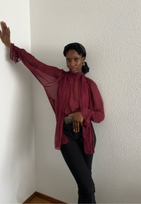 Bordeaux gennemsigtig bluse med lange, flydende ærmer, kombineret med tætsiddende sorte bukser. Modellen bærer et dekorativt bælte og et sort hovedtørklæde.