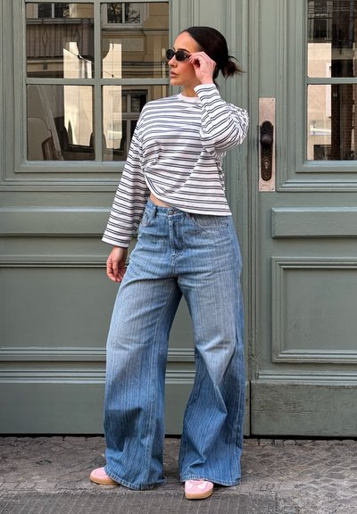Mujer con gafas de sol negras, camiseta de manga larga a rayas, jeans de pierna ancha y zapatillas rosas, de pie frente a unas puertas dobles verdes.