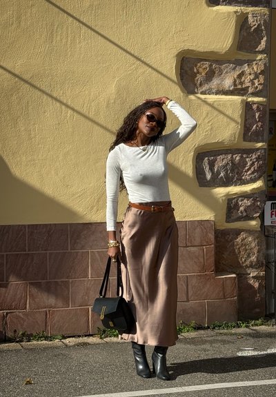 Mujer con gafas de sol que lleva una blusa blanca de manga larga, falda de satén beige, botas negras y sostiene un bolso negro, de pie junto a una pared amarilla.