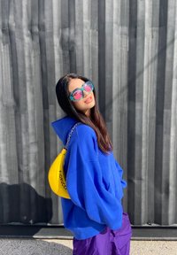 Sweatshirt azul oversized, calças roxas, grandes óculos de sol transparentes e uma mala amarela com a marca "Fiorucci". Fundo texturizado preto.