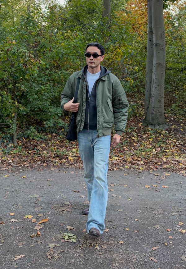 Un hombre con gafas de sol, una chaqueta verde y vaqueros camina por un sendero cubierto de hojas que atraviesa un parque arbolado y lleva un bolso negro al hombro.