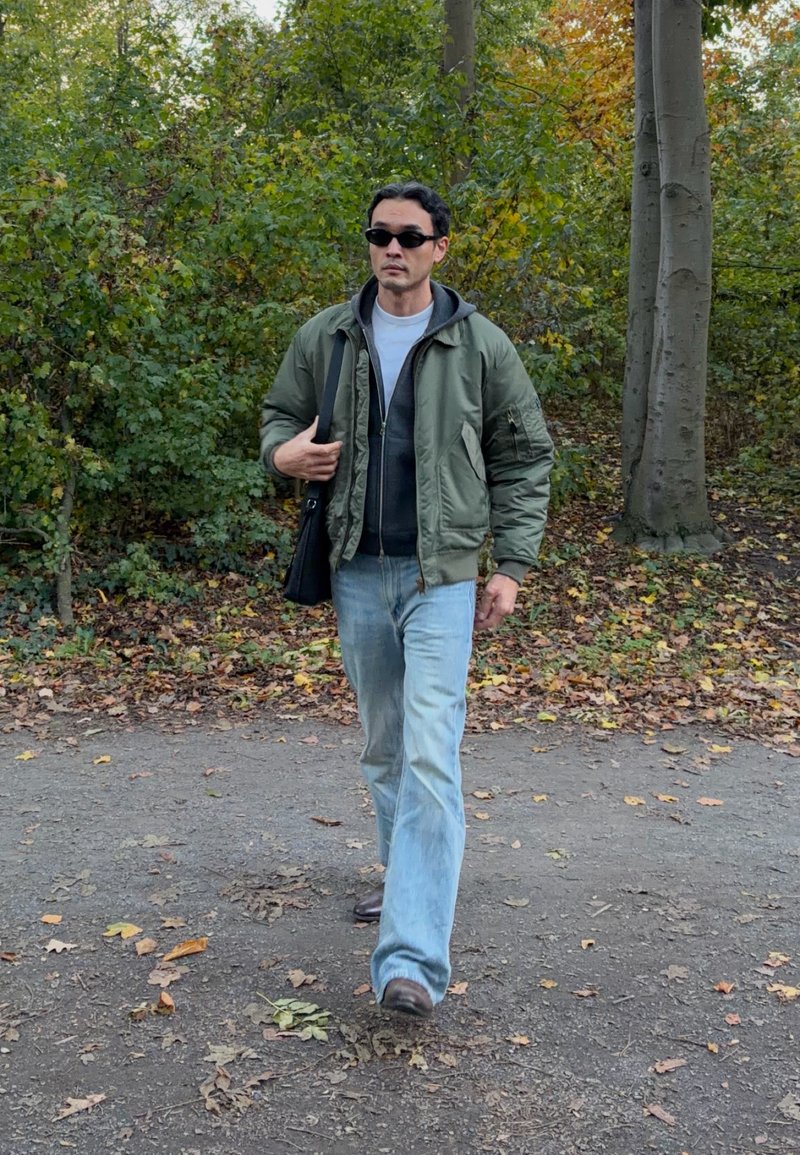 Un homme portant des lunettes de soleil, une veste verte et un jean marche sur un sentier jonché de feuilles dans un parc boisé, avec un sac noir en bandoulière.