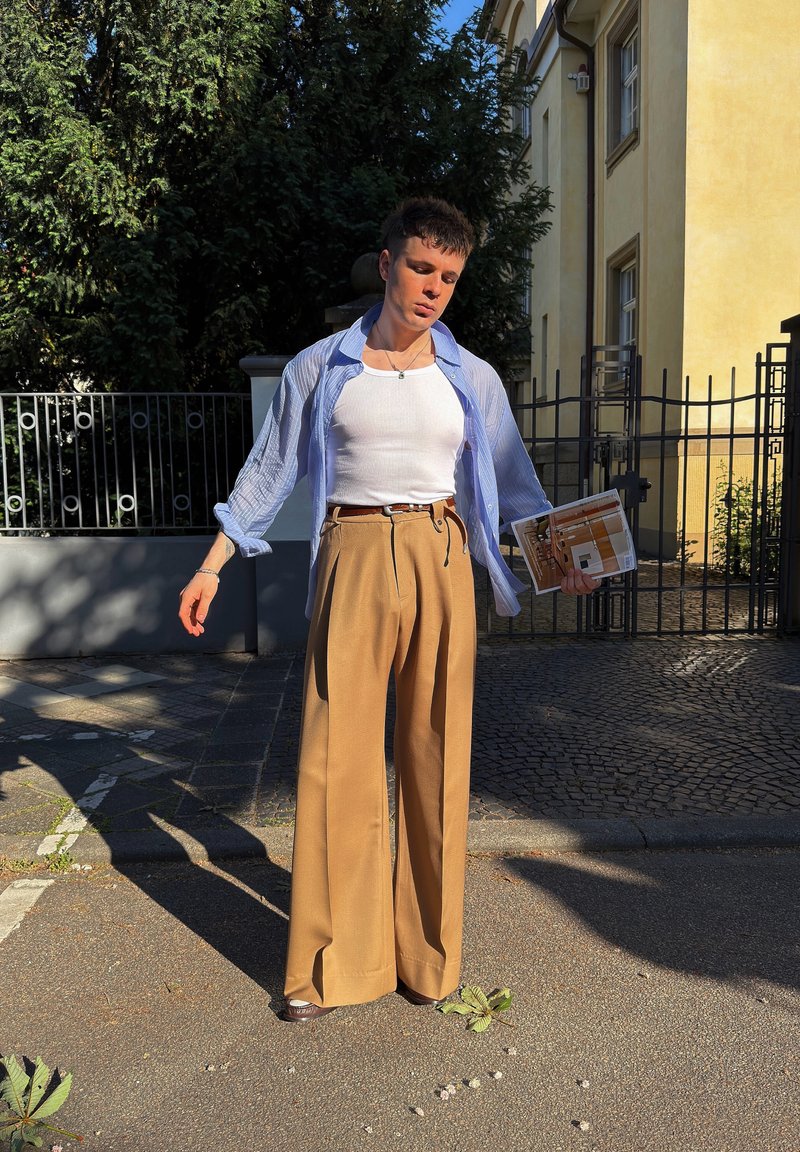 Ein junger Mann trägt weite beige Hose, weißes Tanktop und ein offenes hellblaues Hemd, steht im Freien und hält ein Magazin nahe einem eingezäunten Eingang eines Gebäudes.