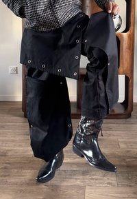 Personne portant un pantalon noir à jambes larges avec boutons-pression et des bottes de cowboy brillantes noires, debout sur un sol en bois à l'intérieur.