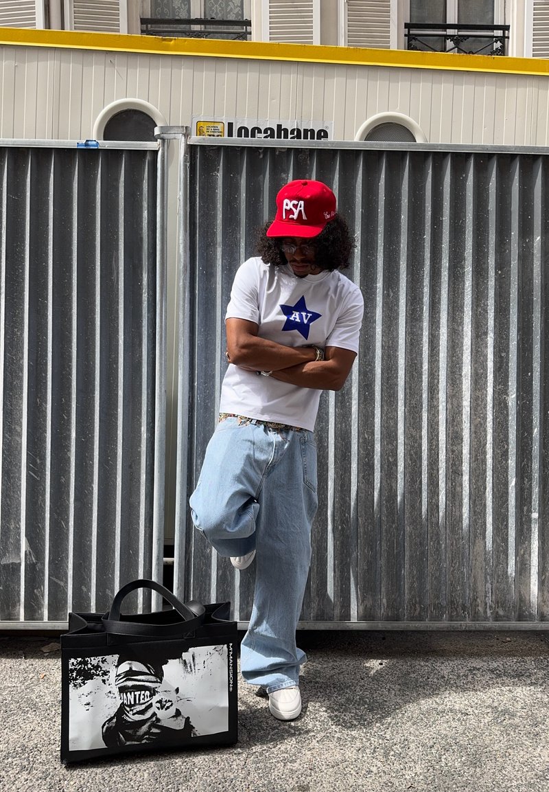 Sac en toile noir avec une impression graphique d'un personnage masqué, associé à un t-shirt blanc arborant une étoile bleue et un jean baggy bleu clair. Casquette rouge.