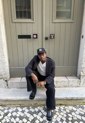Hombre con blazer negro, camisa blanca y gorra negra sentado en un escalón de piedra frente a una puerta doble beige cerrada con buzones y pegatinas.