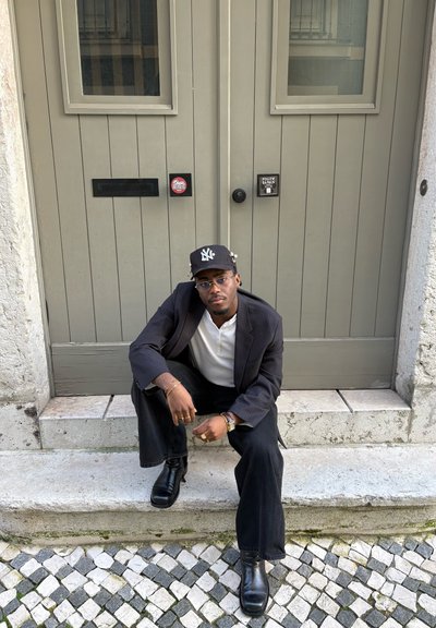 Hombre con blazer negro, camisa blanca y gorra negra sentado en un escalón de piedra frente a una puerta doble beige cerrada con buzones y pegatinas.