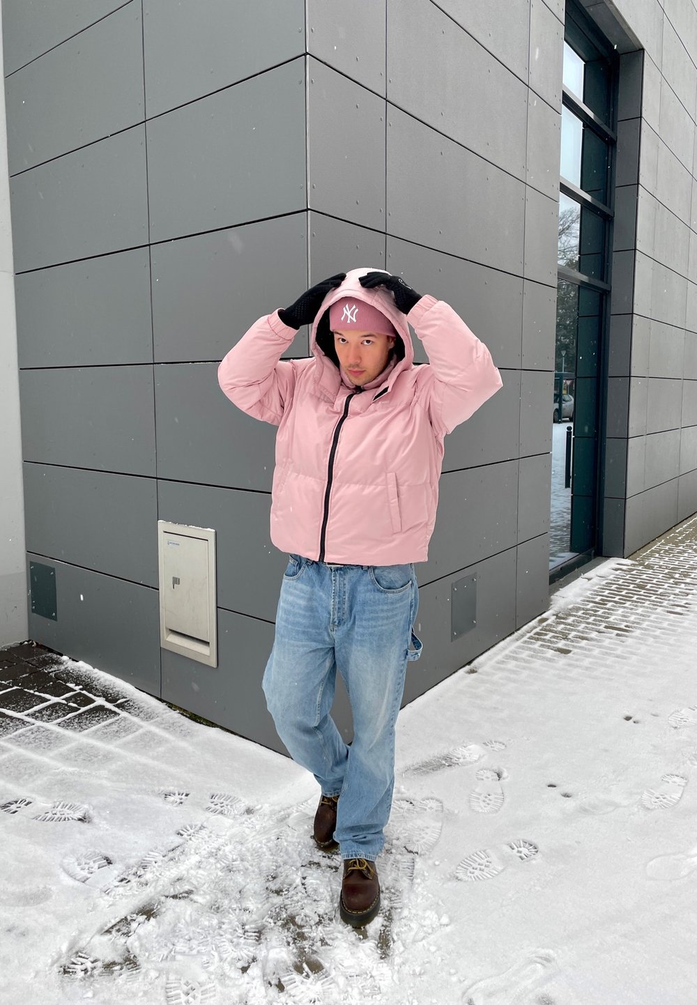 Casaco puffer rosa claro com fecho preto, combinado com jeans azul claro e sapatos castanhos. Chão coberto de neve com pegadas ao fundo.