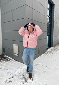 Casaco puffer rosa claro com fecho preto, combinado com jeans azul claro e sapatos castanhos. Chão coberto de neve com pegadas ao fundo.