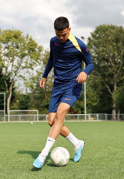 Camiseta deportiva azul de Nike con acentos amarillos, pantalones cortos azules a juego, botas celestes y un balón de fútbol blanco con un patrón sobre césped artificial.