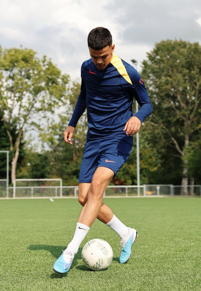 Jersey desportivo azul da Nike com acentos amarelos, calções azuis combinando, chuteiras azul claras e uma bola de futebol branca estampada em relva artificial.
