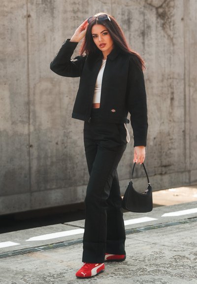 Chaqueta negra corta sobre una blusa blanca, combinada con pantalones acampanados negros y sneakers rojos. Sostiene un pequeño bolso negro. Fondo de concreto texturizado.