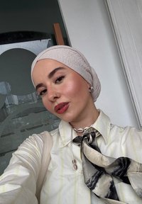 Giovane che indossa un leggero copricapo, orecchini, anello al naso, camicia a righe, collana di conchiglie e una sciarpa attorcigliata nera e beige, all'interno.