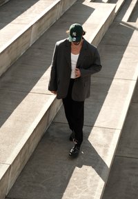 Pelēks pārāk liels žakets pār baltu topu, melnas bikses un melnas kurpes. Zaļa beisbola cepure. Pastaiga pa akmens pakāpieniem, ar ēnām.