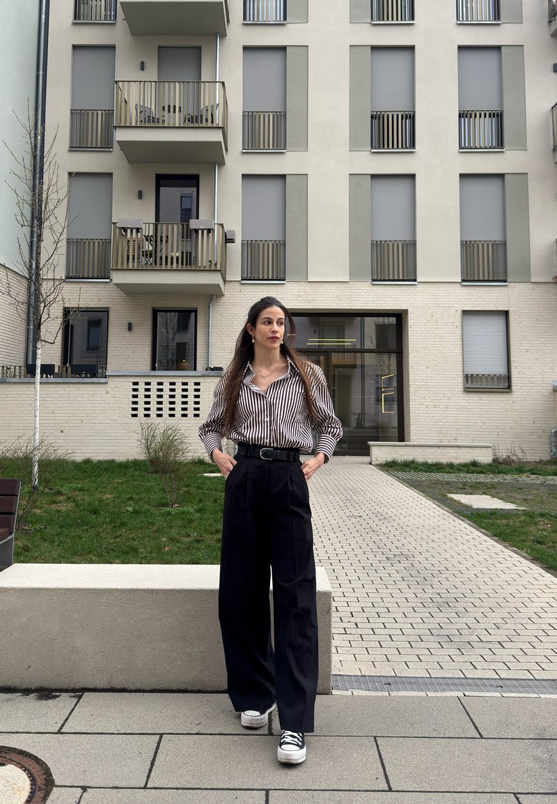 Γυναίκα με μακριά μαλλιά, που φοράει ριγέ πουκάμισο, μαύρο παντελόνι με φαρδιά πόδια και αθλητικά παπούτσια, στέκεται σε ένα μονοπάτι μπροστά από μια πολυκατοικία.