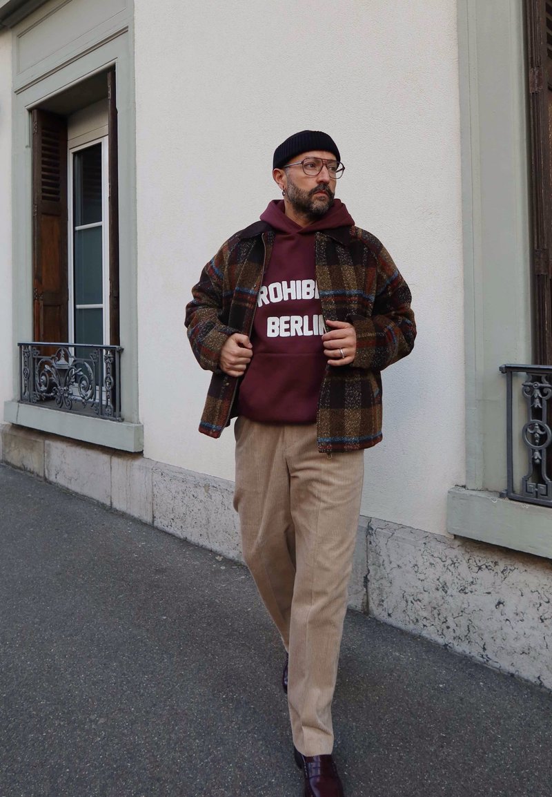 Homme marchant sur le trottoir, portant un bonnet noir, des lunettes, une veste à carreaux, un sweat à capuche bordeaux "PROHIBI BERLIN", un pantalon beige et des chaussures foncées.