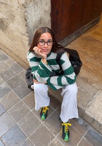 Femme aux longs cheveux bruns et lunettes accroupie sur une rue carrelée, portant une chemise rayée verte et blanche, un pantalon blanc et des baskets noires, vertes et jaunes.