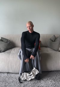 Czarna bluza z długim rękawem na zamek błyskawiczny, szare szerokie spodnie w paski, trzymająca białą torebkę o fakturze z czarnymi akcentami, siedząca na jasnoszarej kanapie.