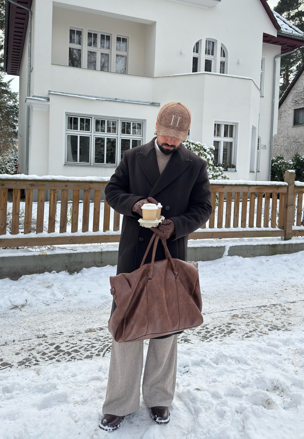 Homme en manteau d'hiver et casquette tenant deux tasses de café et un grand sac en cuir marron, debout dans une rue enneigée devant une maison blanche.