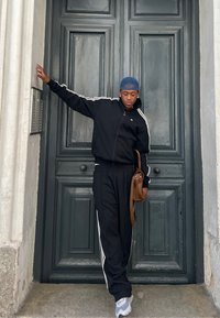 Fekete melegítőruha fehér oldalcsíkokkal, barna táskával párosítva. A téma egy zöld ajtó mellett áll, amelynek textúrázott felülete van. Szürke sportcipő.