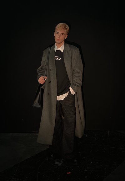 Abrigo gris largo sobre un suéter negro con un logo, camisa blanca de cuello y pantalones de cuadros. Un bolso negro y zapatos completan el atuendo.