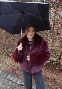 Femme portant un manteau en fausse fourrure violet tenant un parapluie noir, debout sur un chemin pavé avec des buissons feuillus bruns en arrière-plan.