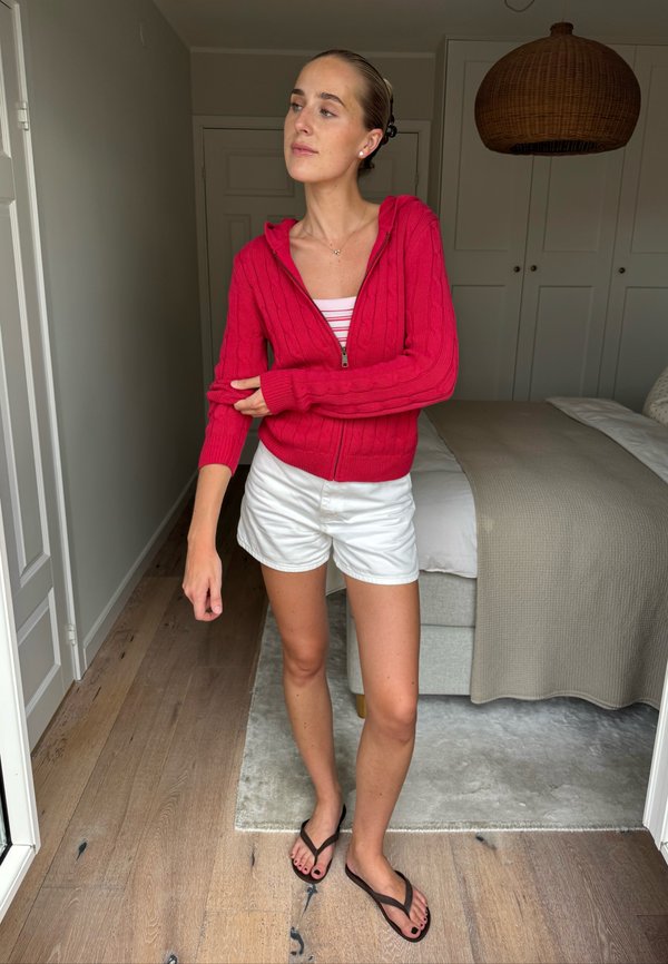 Femme en pull à fermeture éclair rouge, short blanc et tongs noires, debout dans une chambre avec un plancher en bois et une couverture de lit beige.