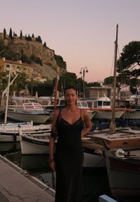 Vestido preto sem mangas com acabamentos em renda, textura suave. Uma mulher está de pé junto a uma marina com barcos, árvores e um penhasco rochoso ao fundo.