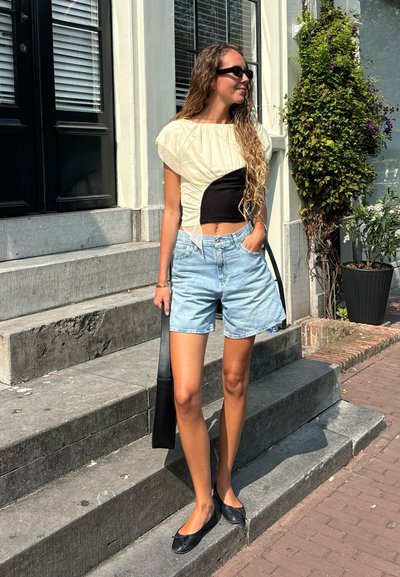 Top crema y negro con mangas abullonadas, combinado con pantalones cortos de mezclilla azul claro. Zapatos planos negros y un bolso negro. Entorno urbano al aire libre.