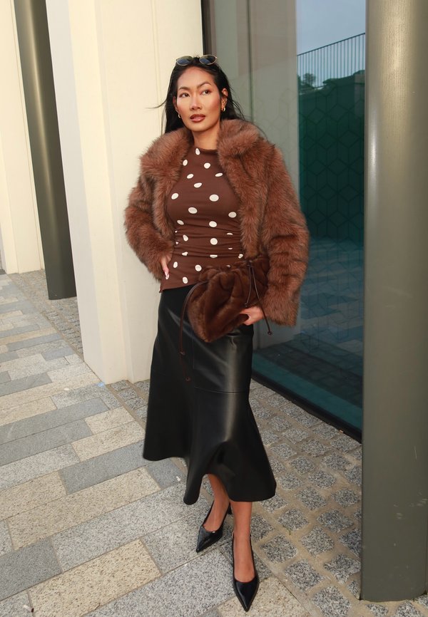 Chaqueta de piel sintética marrón, top marrón con lunares, falda de cuero negra y tacones negros puntiagudos. El conjunto incluye un bolso estructurado y texturas suaves.