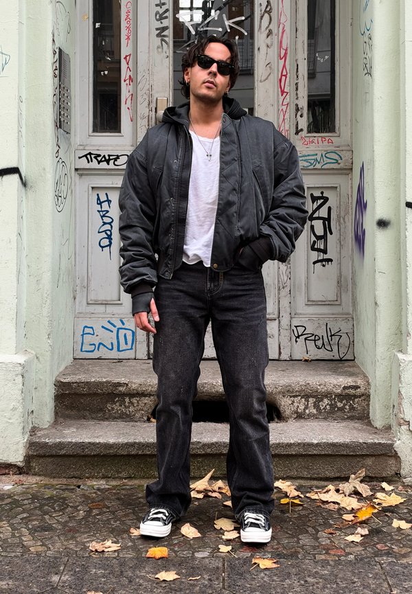 Un homme portant des lunettes de soleil, une veste en cuir noire, une chemise blanche et un jean foncé se tient devant des portes couvertes de graffitis, les mains dans les poches.