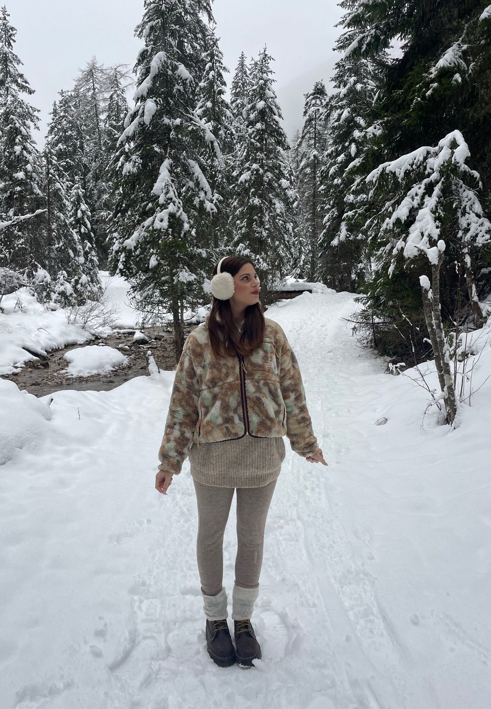 Fleecejacke mit braunem und grünem Muster, beiges Sweatshirt und beige Leggings mit Fellabschlüssen. Dunkle Stiefel. Verschneiter Wald im Hintergrund.