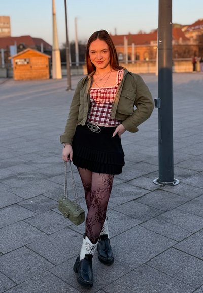 Mujer joven está de pie al aire libre en una zona pavimentada, lleva puesto un top de cuadros, chaqueta color oliva, falda negra, medias con estampado y botas en blanco y negro, sosteniendo un bolso verde.