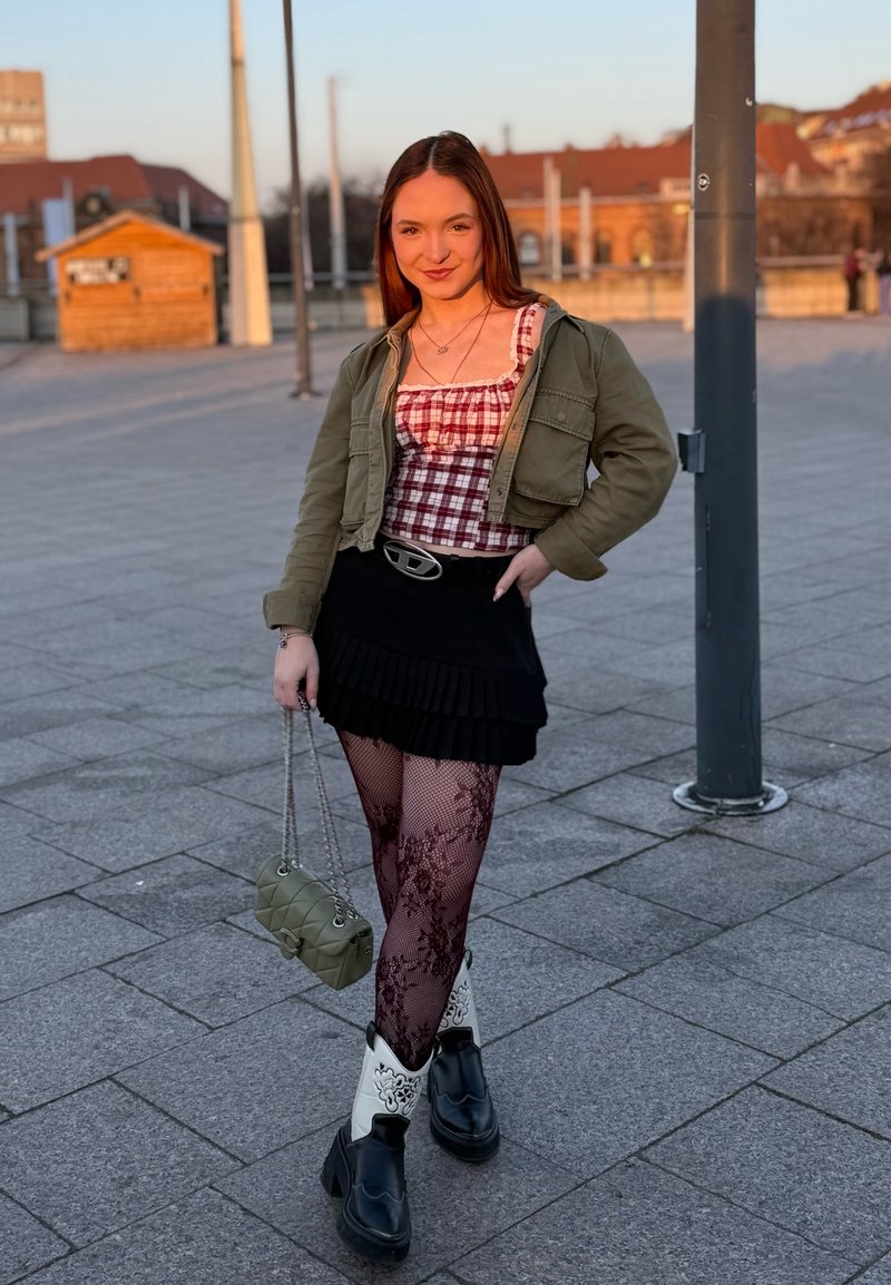 Jeune femme debout dehors sur une zone pavée, portant un haut à carreaux, une veste olive, une jupe noire, des collants à motifs et des bottes noires et blanches, tenant un sac vert.