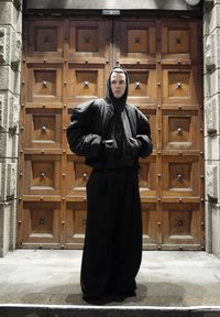 Juoda per didelis pūstas paltas su gaubtu, sluoksniuotas ant juodo džemperio, derinamas su laisvomis juodomis kelnėmis. Medinės durys fone su žvaigždžių detalėmis.