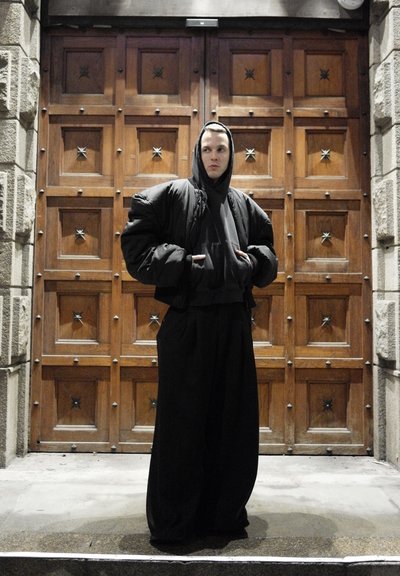 Chaqueta negra extragrande y acolchada con capucha, combinada con una sudadera negra y pantalones negros holgados. Fondo de una puerta de madera con detalles de estrellas.