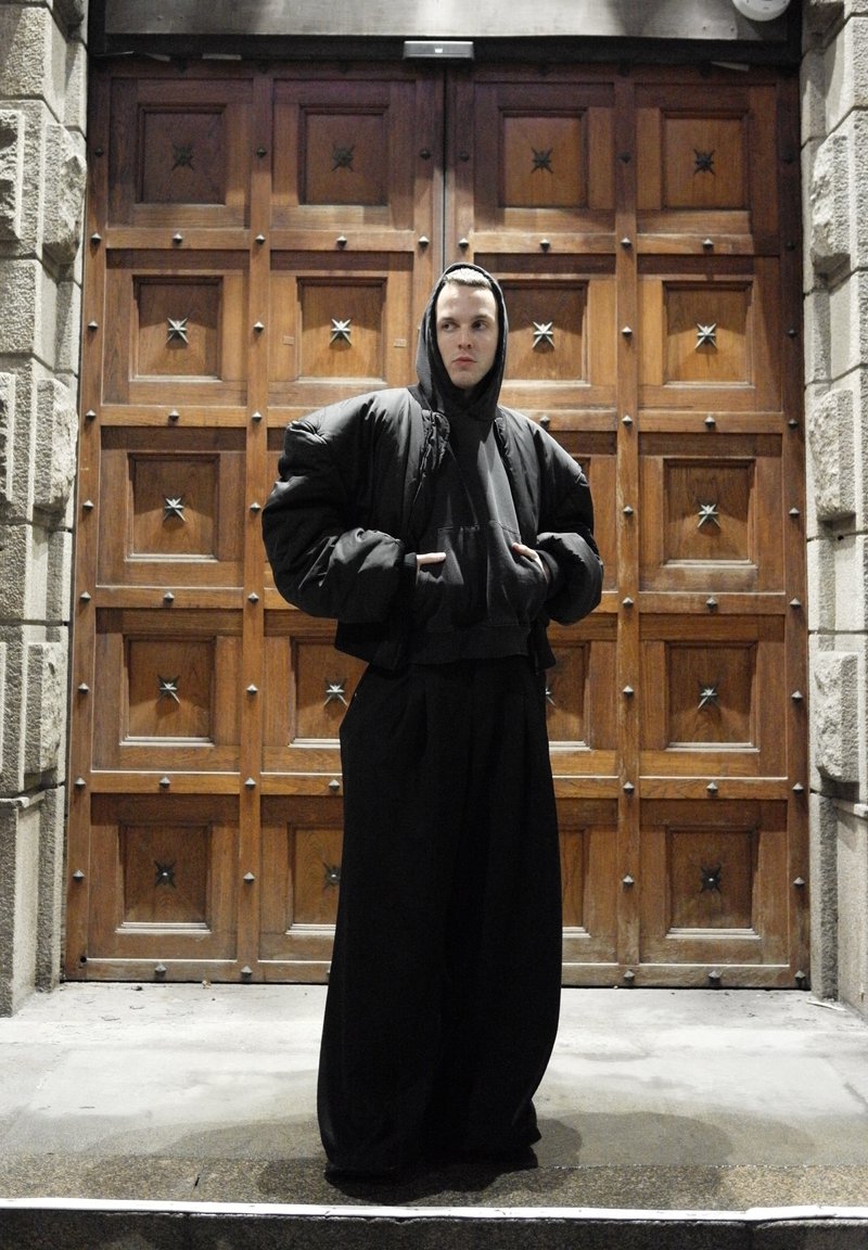 Schwarze, übergroße, wattierte Jacke mit Kapuze, darüber ein schwarzer Pullover, kombiniert mit locker sitzenden schwarzen Hosen. Holztür im Hintergrund mit Sternverzierungen.